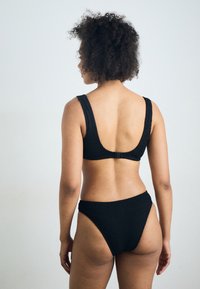 Czarny bikini z prążkowanego materiału, z szerokimi ramiączkami i niskim krojem pleców. Wysokie wcięcia na dole. Teksturowany materiał i czyste linie podkreślają nowoczesny wygląd.