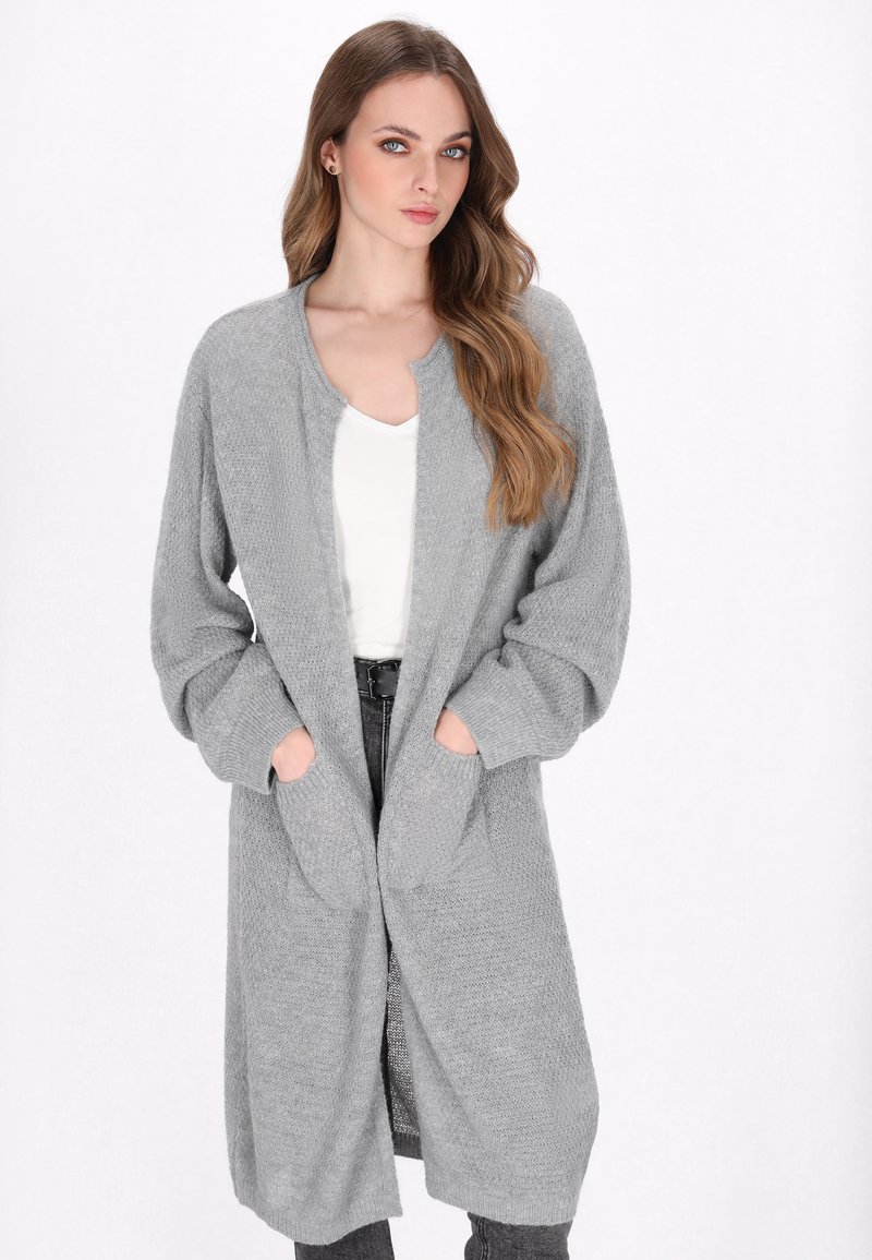 Cardigan lung, tricotat, de culoare gri deschis, cu o suprafață texturată, cu fața deschisă, două buzunare frontale și mâneci largi. Purtați peste un top alb.