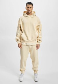 Beige oversized hoodie med en kengurulomme, parret med matchende joggerbukser. Stoffet ser blødt ud, med elastiske manchetter og talje.