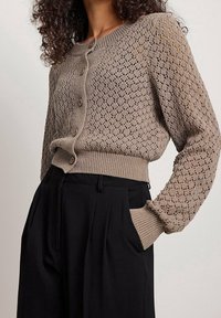 Beige neulecardigan avoimella kuviolla, pyöreällä pääntiellä ja nappisuljennalla. Varustettu ribbireunuksilla hihoissa ja helmassa, käytetään korkeavyötäröisten mustien housujen kanssa.