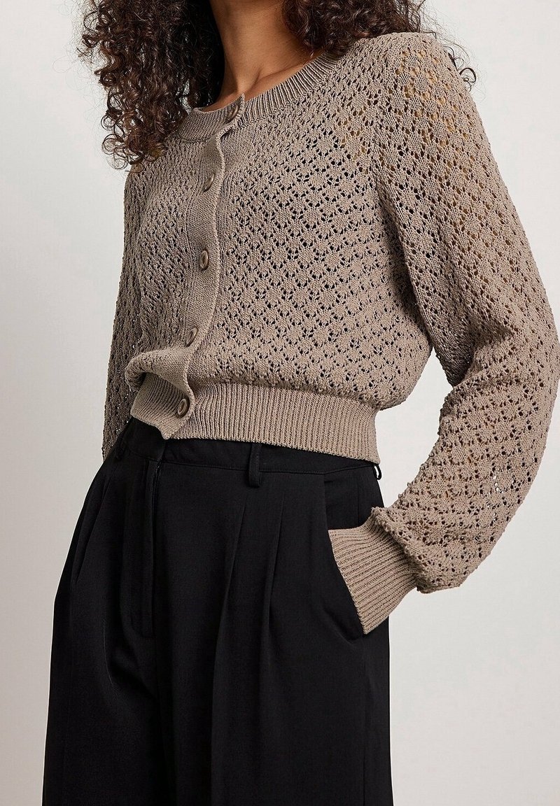 Beige neulecardigan avoimella kuviolla, pyöreällä pääntiellä ja nappisuljennalla. Varustettu ribbireunuksilla hihoissa ja helmassa, käytetään korkeavyötäröisten mustien housujen kanssa.