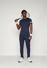 Merenkantoinen urheiluvaatetus, joka sisältää istuvan t-paidan, jonka hihassa on logo, ja yhteensopivat trikoot. Asusteena valkoiset lenkkarit ja tennismaila.