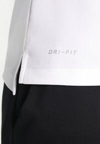 Tessuto bianco con la scritta "DRI-FIT" sopra un tessuto nero, mostrando le cuciture e una piccola apertura laterale sul materiale bianco.