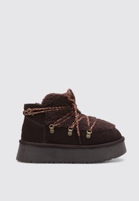 Bota de ante marrón con un cuello de forro polar texturizado, con cordones trenzados multicolores y herrajes en tono dorado. Suela gruesa y acolchada.