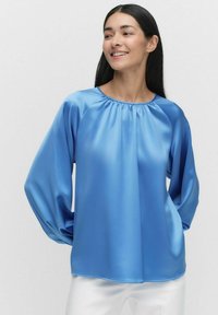 Blå satinblus med rund halsringning och skrynkliga detaljer. Har långa, lösa ärmar och en mjuk, glansig yta.