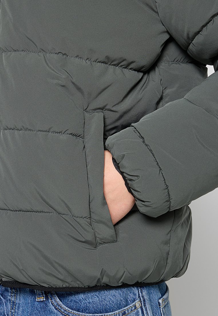 Chaqueta acolchada de color gris oscuro con un diseño de rombos, que cuenta con un bolsillo lateral y puños elásticos, llevada sobre pantalones de mezclilla. Textura suave.