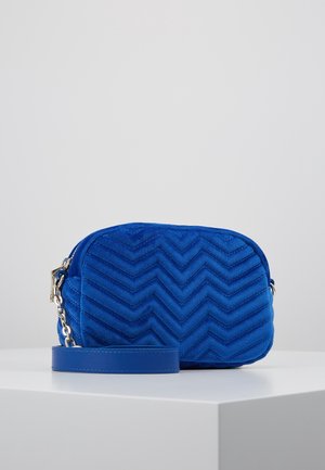 Mała prostokątna torebka na ramię z pikowanego aksamitnego materiału w kolorze królewskiego błękitu, z wzorem w kształcie szewronu, złotym zamkiem błyskawicznym oraz dopasowanym niebieskim skórzanym paskiem, na białym tle.