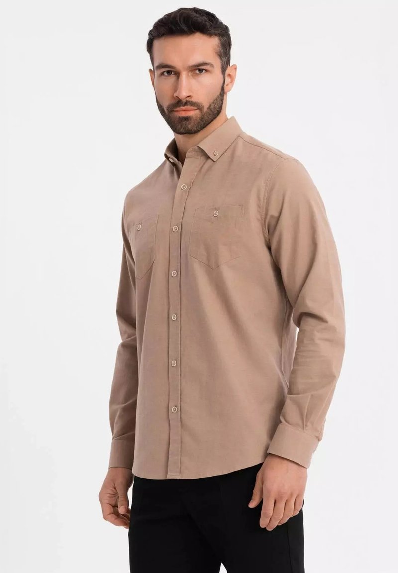 Beige Langarmhemd mit Button-Down-Kragen, zwei Brusttaschen und strukturiertem Stoff. Kombiniert mit schwarzen Hosen.