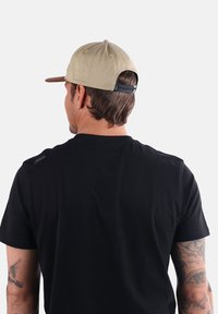 Casquette de baseball kaki avec une visière plate, strap ajustable à l'arrière, portée par une personne en t-shirt noir, montrant un design et une texture simples.