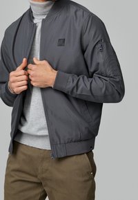 Grijze bomberjack met een gladde textuur, geribde manchetten en een ritszak op de mouw. Draag het over een lichtgrijze coltrui.