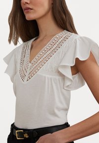 White short-sleeve top with a deep V-neck, featuring intricate lace detailing and ruffled sleeves. Smooth fabric against a black belt.