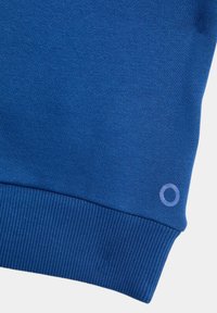 Cuff di felpa blu con texture a costine, tessuto liscio e un sottile logo circolare di colore contrastante sul lato.