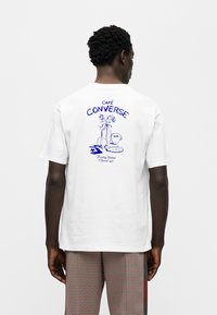 Λευκό βαμβακερό T-shirt με μπλε γραφικό μιας κούπας καφέ και λουλουδιών στην πλάτη, με το κείμενο "CAFÉ CONVERSE" από πάνω.