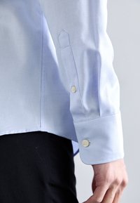 Svetlomodrá textúrovaná košeľa s krémovými gombíkmi na manžetách, nosená s čiernymi nohavicami, s uvoľnenou rukou pri boku na jednoduchom pozadí.