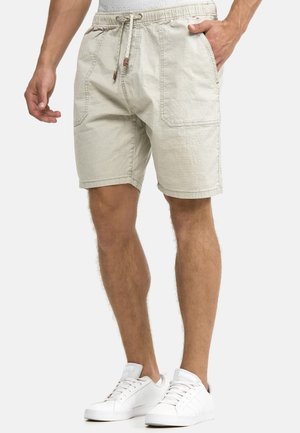 Mann trägt hellbeige Shorts mit Kordelzug und Fronttaschen und weiße Sneaker, steht vor einem einfarbigen Hintergrund.