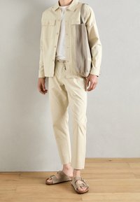Beżowa bawełniana kurtka z dwoma przednimi kieszeniami, zestawiona z dopasowanymi spodniami. Wykończona jasnoszarymi sandałami i szarym płóciennym torbą.