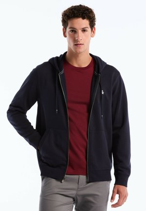 Marineblauer Reißverschluss-Hoodie mit einer Brusttasche, Kordelzugkapuze und kontrastierenden Nähten, kombiniert mit einem weinroten T-Shirt und grauen Hosen.