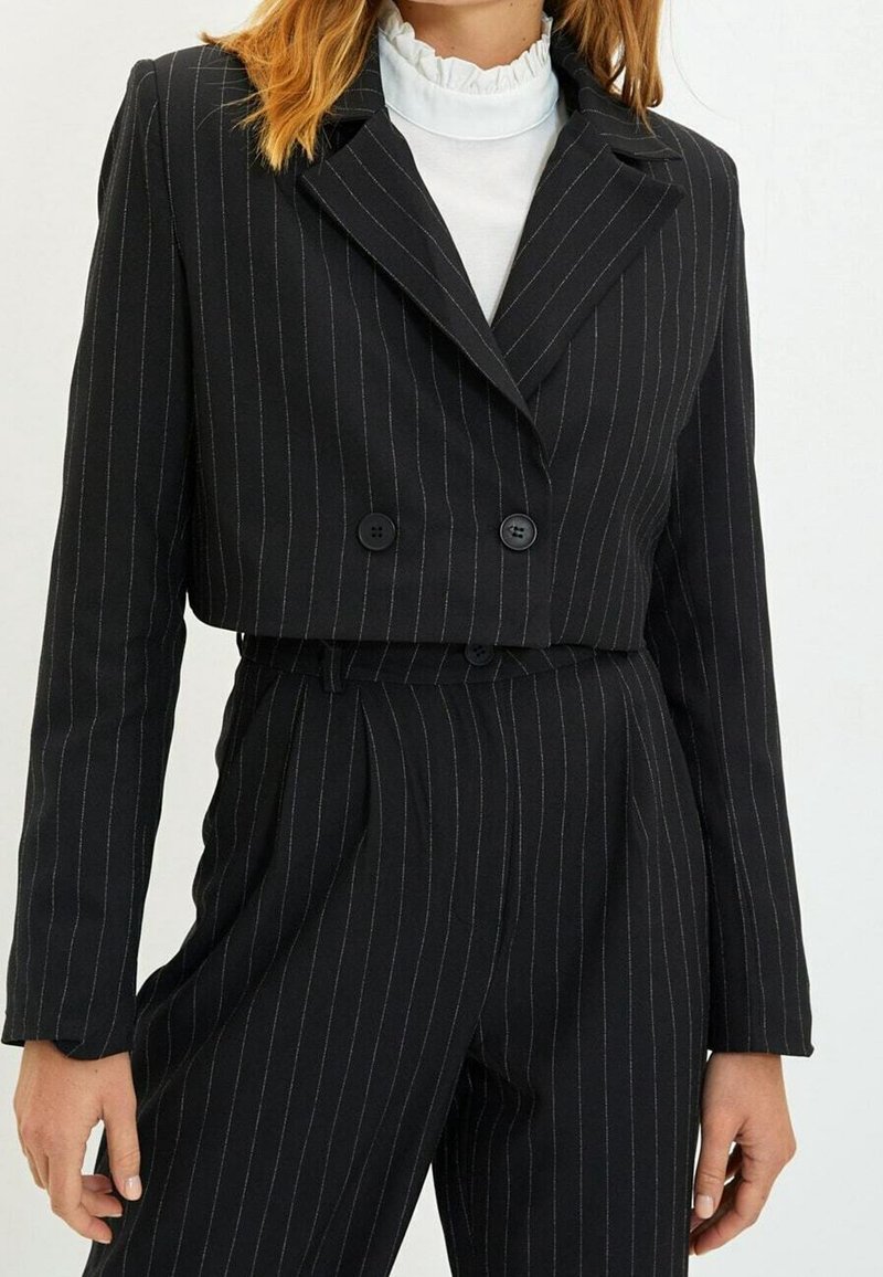 Femme portant un blazer noir à fines rayures coupé, à double boutonnage, sur un pantalon taille haute assorti et un chemisier blanc à col volanté.