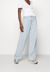 Jeans anchos de color azul claro con cintura alta, combinados con una blusa blanca ajustada con escote en forma de V y dobladillo con volantes. Zapatillas deportivas plateadas completan el look.