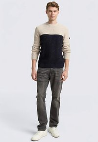 Strickpullover mit einem beigen Oberteil und einem dunkelblauen Unterteil, ausgestattet mit einer gerippten Textur und gesprenkelten Details. Kombiniert mit grauer Jeans.