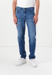 Vaqueros de mezclilla azul con un corte recto, diseño de cinco bolsillos, trabillas para el cinturón y un ligero desvanecimiento. Combinados con zapatillas blancas.
