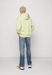 Sudadera verde claro con capucha, de corte holgado, combinada con pantalones vaqueros azules deslavados y zapatillas multicolores. La textura parece suave y casual.