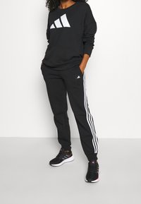 Pantalones de chándal negros con rayas blancas laterales, combinados con un sudadera negra que tiene un gran logo blanco y zapatillas deportivas negras.