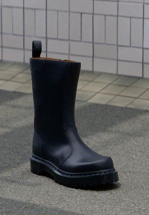 Black leather mid-calf boot with pull tab, chunky lug sole, and smooth rounded toe, standing on paved ground with tiled wall background.