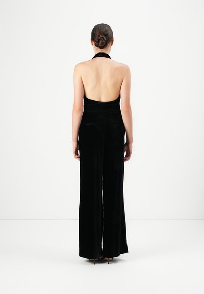 Vrouw met vastgebonden haar, gekleed in een zwart fluwelen halter-jumpsuit en hakken, staande met haar gezicht naar een witte achtergrond.