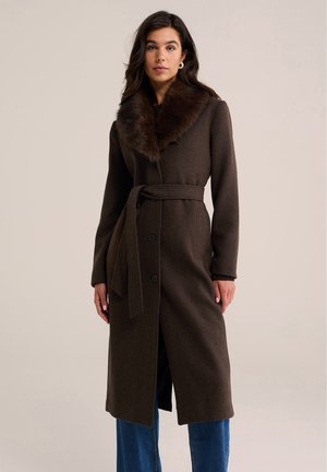 Femme portant un long manteau marron foncé avec un col en fourrure, une taille cintrée avec ceinture, une fermeture à boutons et un jean bleu, debout devant un fond uni.