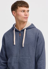 Blue zip-up hoodie with beige drawstrings, featuring a soft texture and front pockets. Includes a contrasting inner lining.