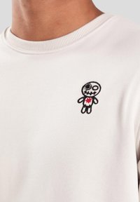 Sudadera blanca con un pequeño personaje bordado en negro y rojo en el pecho, cuello redondo con detalles acanalados.