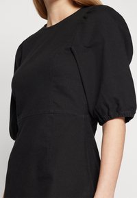 Czarna sukienka z krótkimi, bufiastymi rękawami, okrągłym dekoltem i dopasowanym korpusem. Wykonana z gładkiego materiału z delikatnymi szwami.