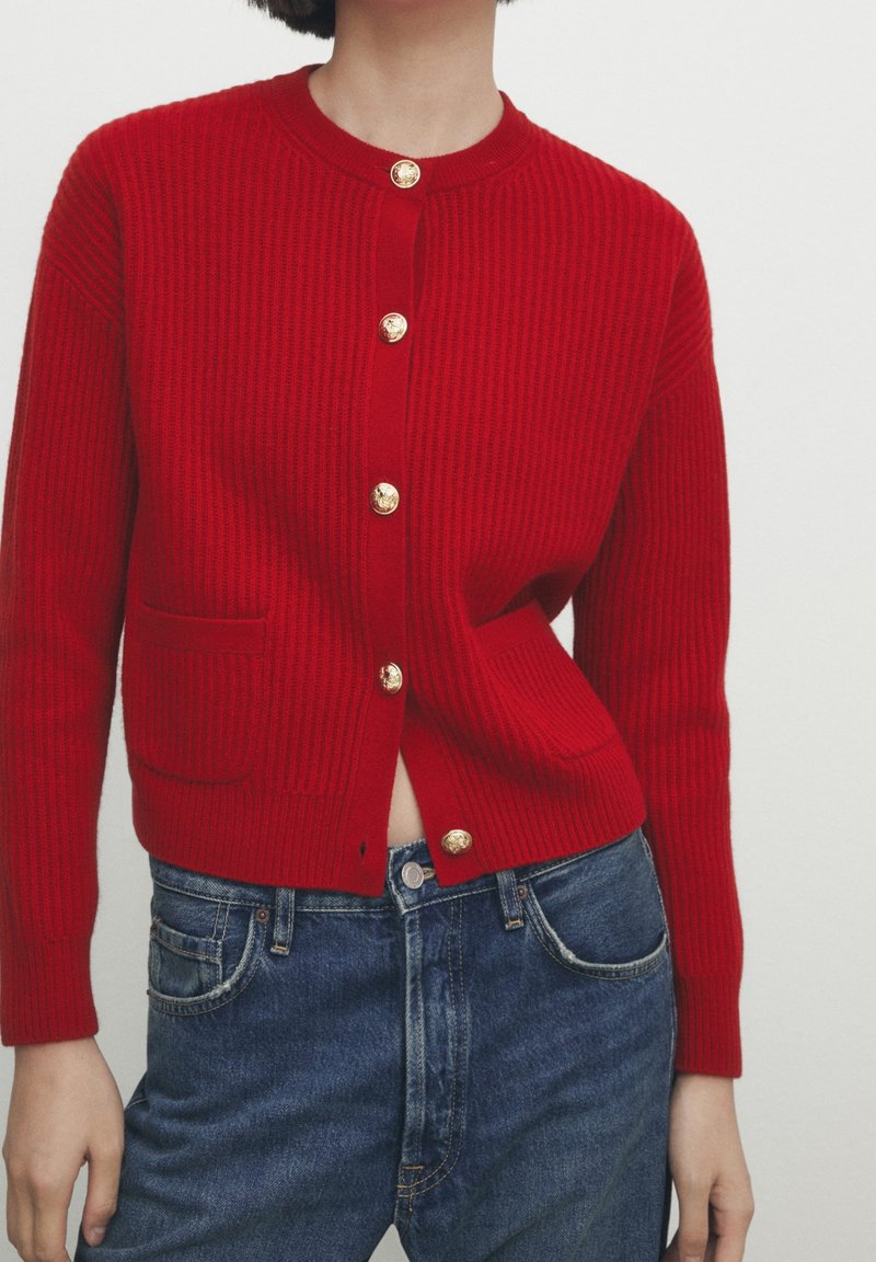 Femme portant un cardigan côtelé rouge avec des boutons dorés et deux poches avant, associé à un jean taille haute bleu.