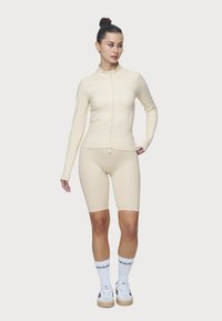 Beige cipzáros dzseki és hozzá illő biciklis sort sima anyagból. A modellen fehér zokni van fekete logóval és fehér sportcipő.