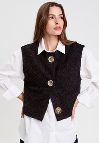 Schwarwest textured Weste mit drei goldenen Knöpfen, die über einem weißen Hemd getragen wird. Sie hat ein kurzes Design mit offenem Vorderteil und breiten Schultern.