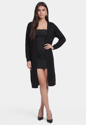 Kvinde stående iført en sort stropløs minikjole med en lang sort åben cardigan, sorte hæle og store guld hoop øreringe.