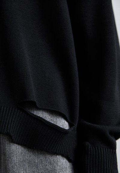 Pull en maille noir avec un design côtelé texturé et un détail découpé distinct au niveau de l'ourlet, superposé sur un tissu gris.