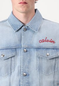Vaaleansininen farkkutakki, jossa on kaksi rintataskua, hopeiset napit ja punaisella brodeerattu "calvin" oikeassa yläkulmassa.