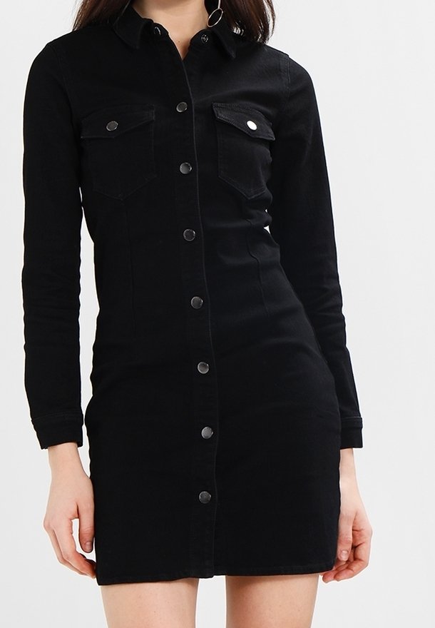 Femme portant une robe en jean noire cintrée à boutons, manches longues et deux poches poitrine avec rabats et boutons.
