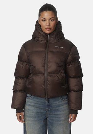 Mujer con cabello oscuro lleva una chaqueta acolchada marrón con capucha y cremallera, combinada con vaqueros azules, de pie frente a un fondo sencillo.