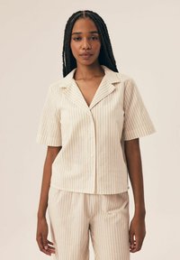 Koszula z krótkim rękawem i kołnierzykiem w jasnym beżu z cienkimi pionowymi paskami, wykonana z materiału o miękkiej fakturze, zestawiona z podobnymi spodniami w paski.