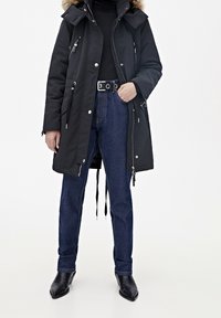 Czarna parka z kapturem wykończonym futerkiem, z zamkami błyskawicznymi i kieszeniami, zestawiona z niebieskimi, wysokim stanem dżinsami oraz czarnymi botkami na obcasie.