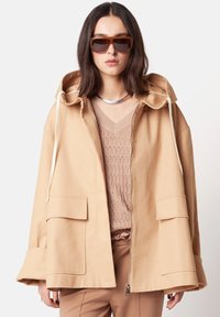 Beige Kapuzenjacke mit silbernem Reißverschluss, zwei Fronttaschen und Kordelzügen, getragen über einem strukturierten hellen Oberteil, kombiniert mit braunen Sonnenbrillen.
