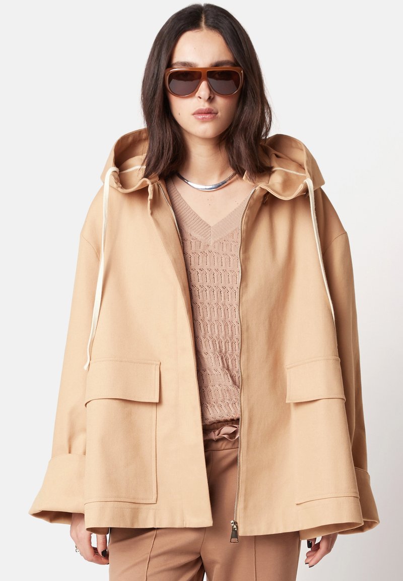 Beige Kapuzenjacke mit silbernem Reißverschluss, zwei Fronttaschen und Kordelzügen, getragen über einem strukturierten hellen Oberteil, kombiniert mit braunen Sonnenbrillen.