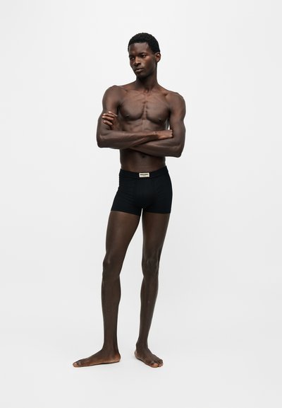 Homme torse nu, debout pieds nus, les bras croisés, portant un boxer noir sur un fond clair uni.
