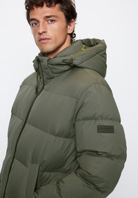 Padded green jacket with a hood, featuring a zip closure, large front pockets, and a fabric logo patch on the sleeve. Textured surface.