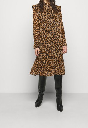 Femme portant une robe mi-longue à manches longues imprimé léopard avec un ourlet à volants et des bottes noires montantes jusqu'aux genoux sur un fond uni.