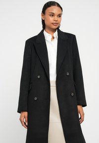 Casaco de mistura de lã preto com fecho de botões duplos, lapela recortada e bolsos laterais. Textura suave, design até ao joelho.