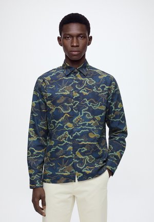Homme portant une chemise bleu marine boutonnée avec un motif abstrait vert et jaune rappelant une carte et un pantalon blanc cassé, debout devant un fond blanc.